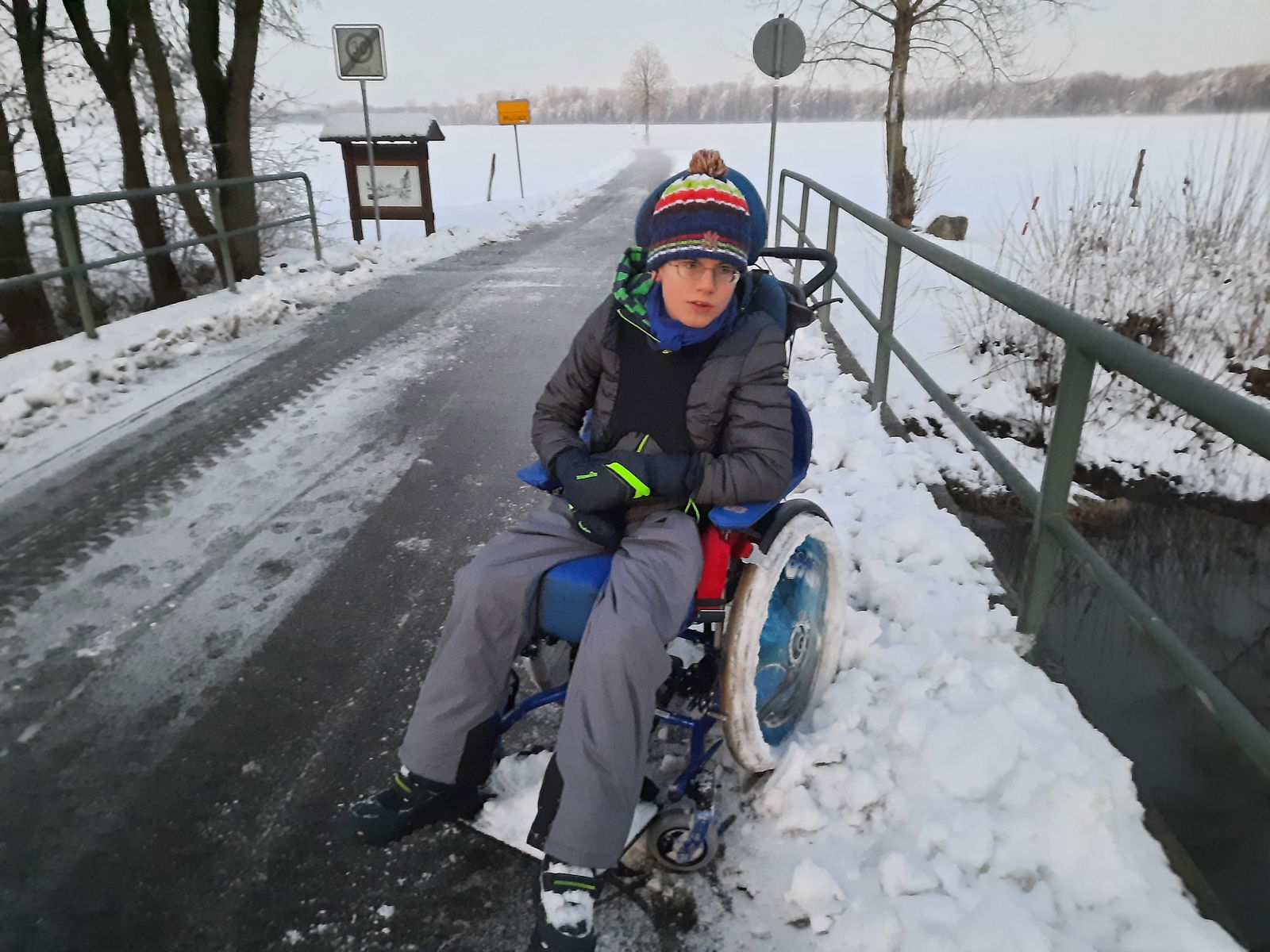 Jugendlicher mit Mütze und dicker Winterjacke im Rollstuhl auf schneebedeckter Straße