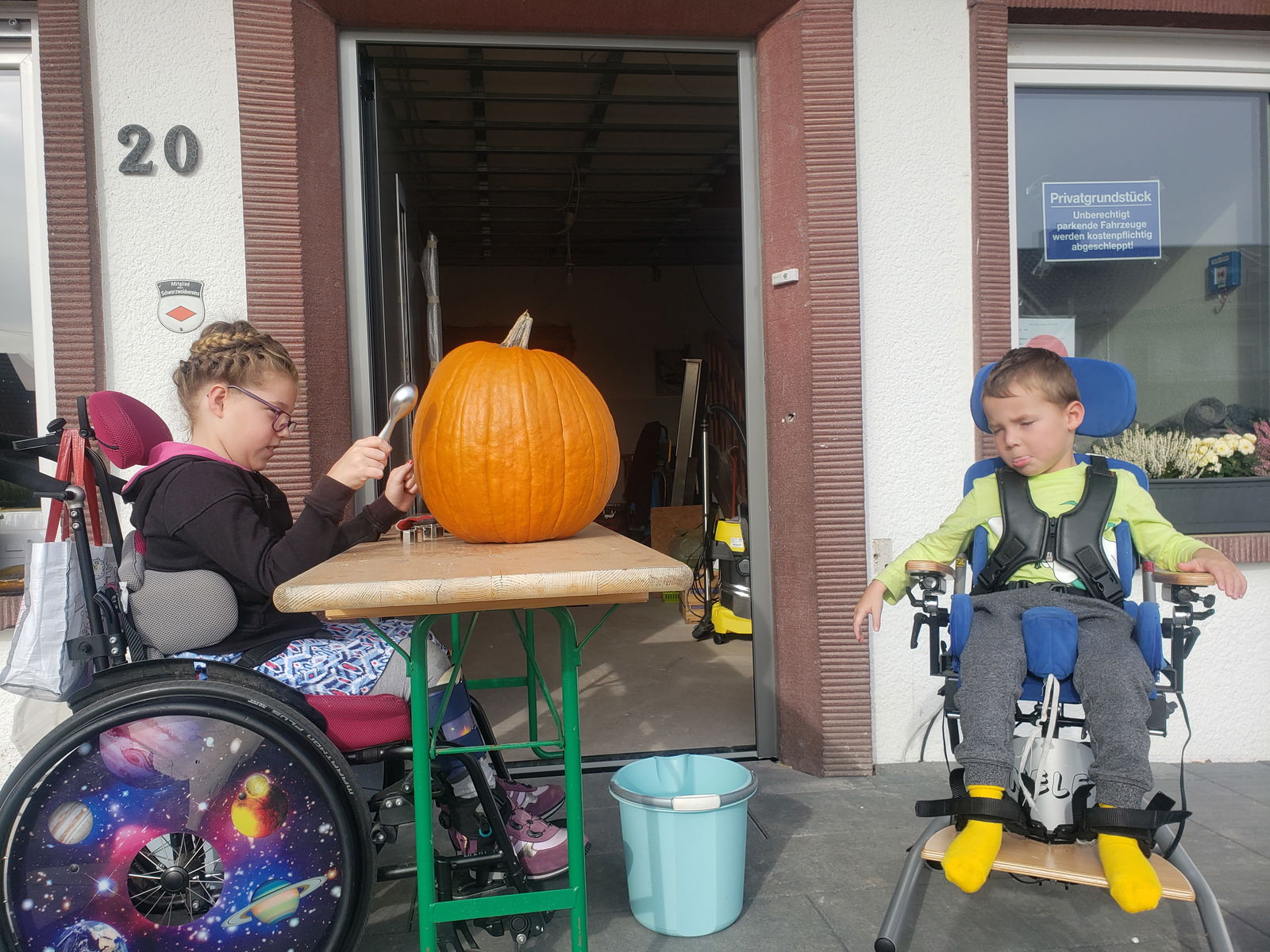 Kleines Mädchen sitzt im Rollstuhl und arbeitet am Kürbis; kleiner Junge sitzt daneben im Rollstuhl