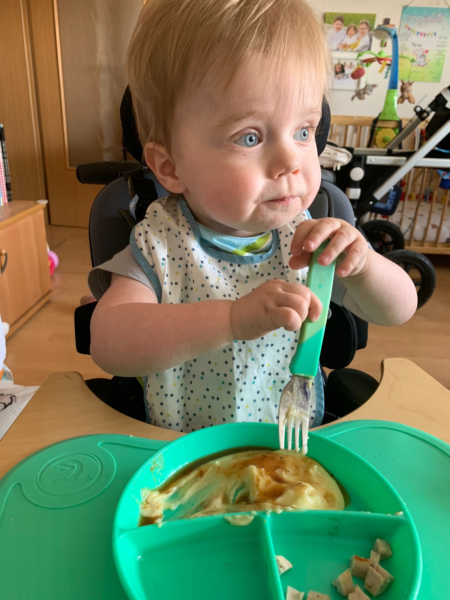 Kleinkind auf speziellem Kindersitz mit Gabel und Brei und schaut mit großen Augen 