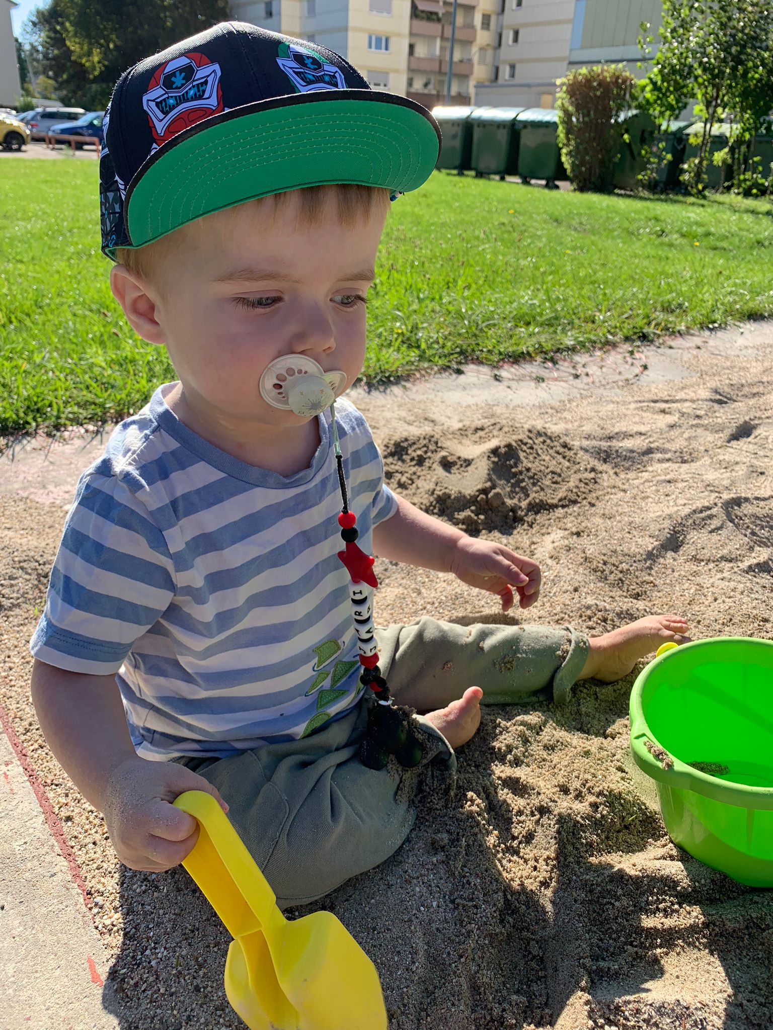 Junge mit Mütze, Schnuller, Schaufel und Eimer im Sand