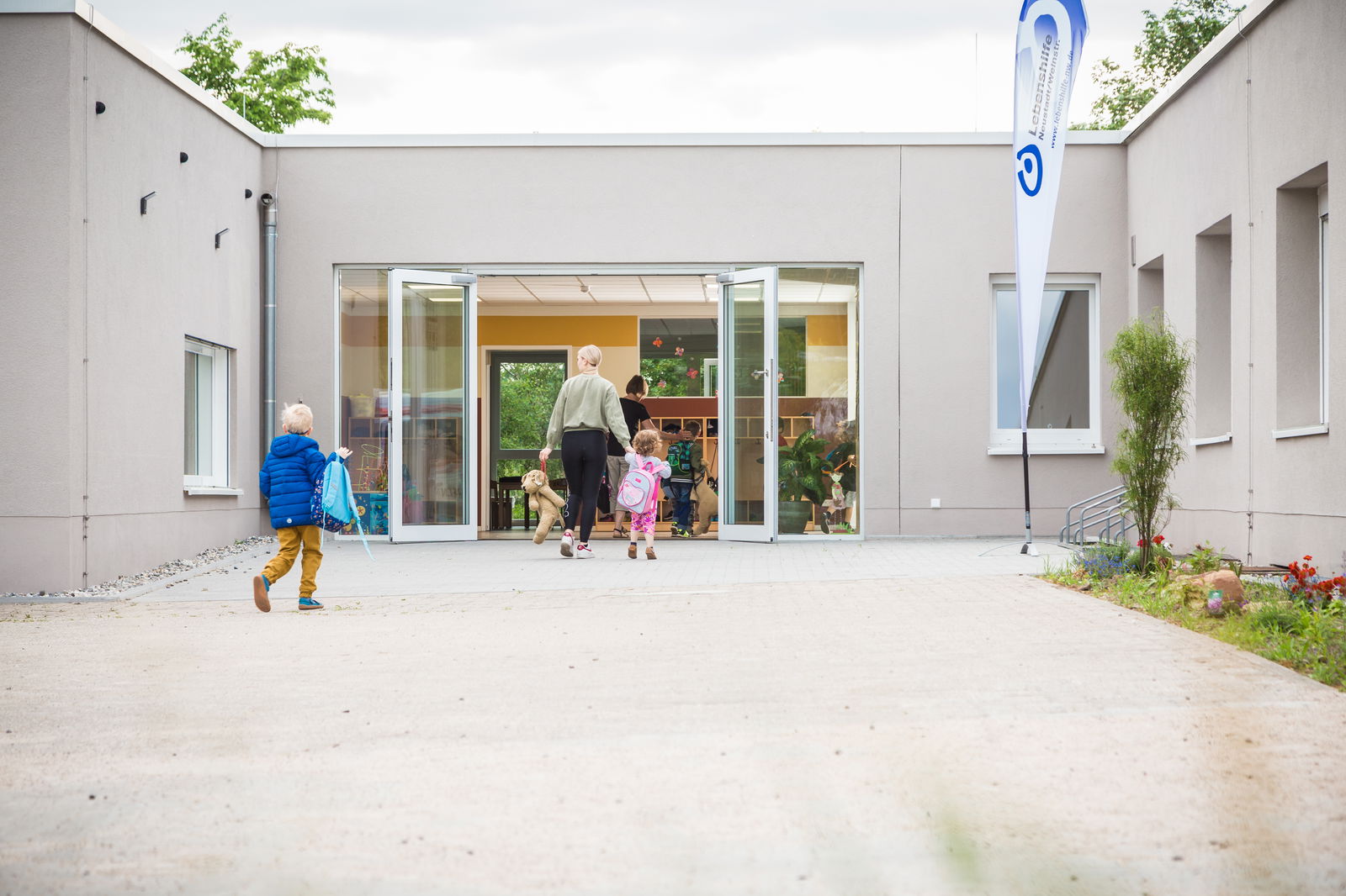 Mutter mit Tochter und Teddy auf dem Weg zur Kita, Junge mit Rucksack kommt hinterher