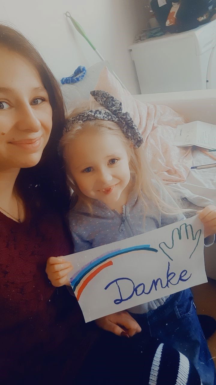 Lächelndes Mädchen im Arm ihrer Mutter hält ein Danke-Schild hoch auf dem auch ein Regenbogen mit einer Hand gemalt ist