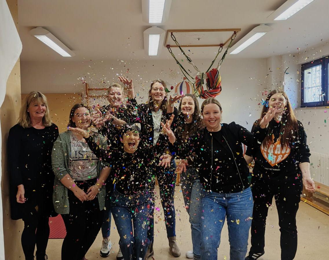 Mehrere Menschen stehen in der Turnhalle, lachen, winken und werfen Konfetti