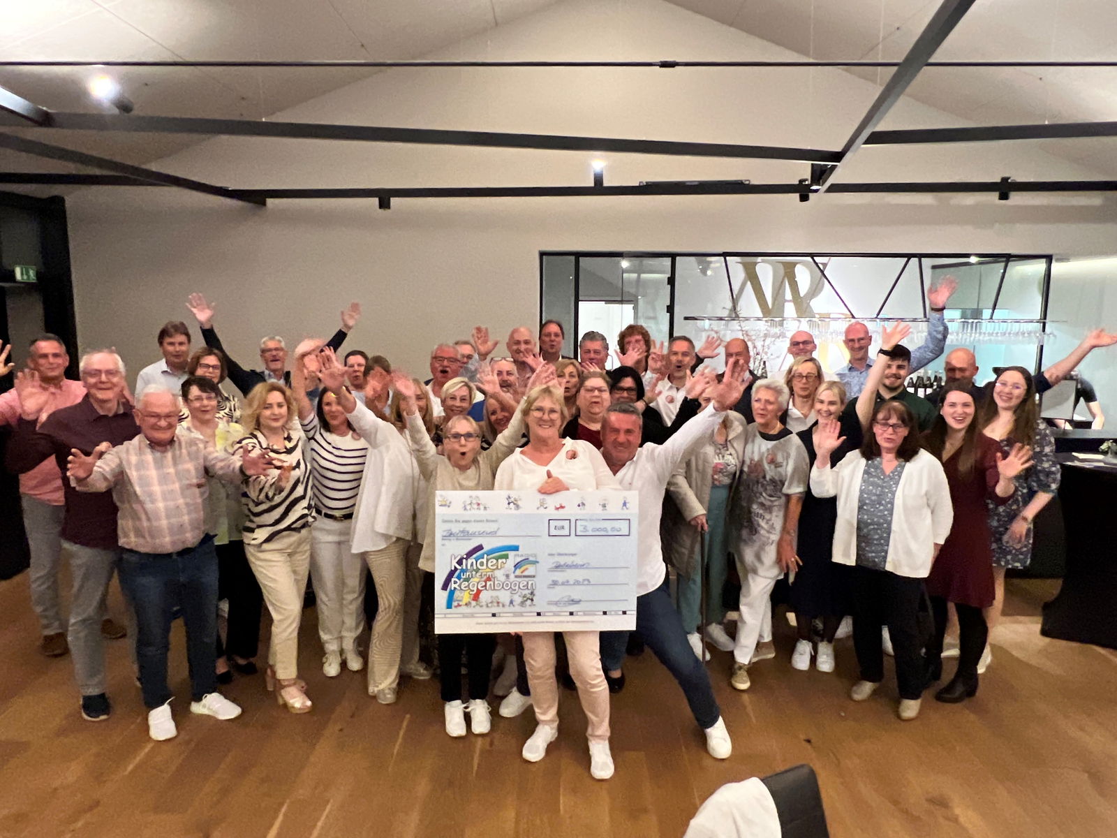 Frau Bätz mit Scheck in der Hand für Kinder unterm Regenbogen mit feiernden Menschen im Hintergrund