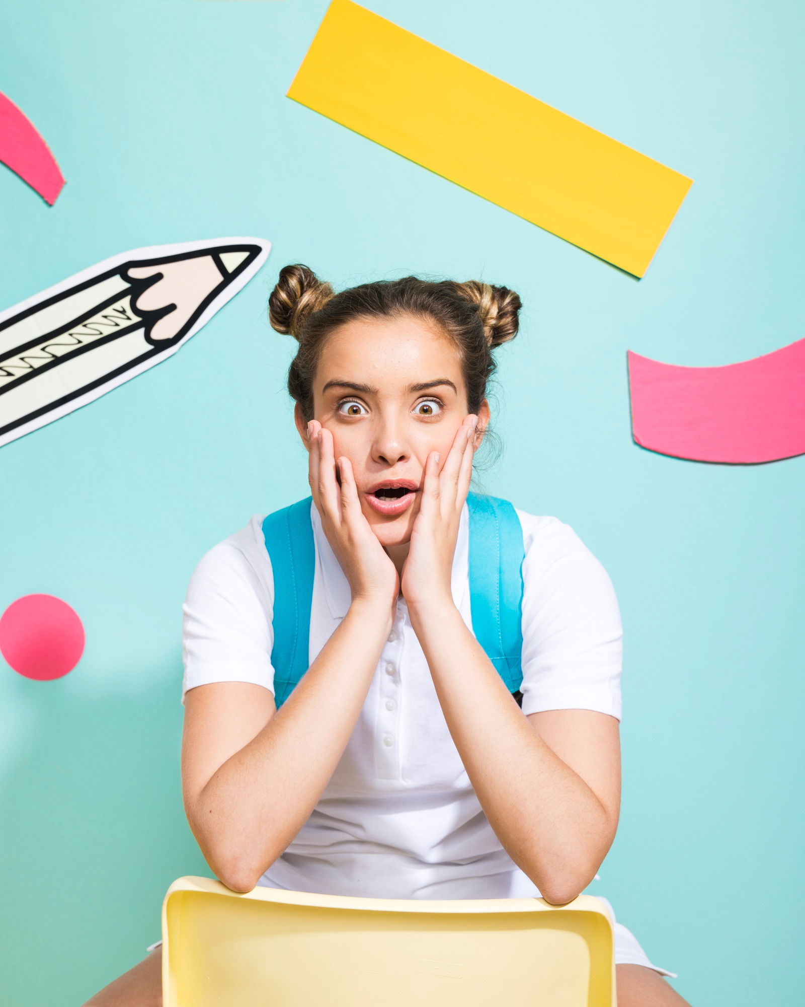 Eine Schülerin mit Zöpfen sitzt vor einem bunten Hintergrund und trägt einen blauen Schulranzen. Sie hält sich schockiert blickend bei offenem Mund die Hände ins Gesicht 