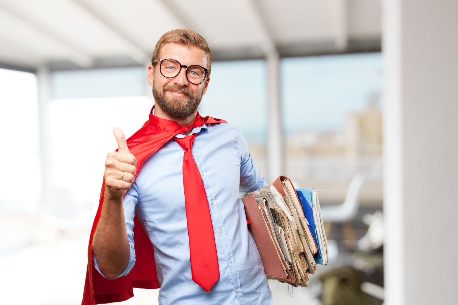 Ein blonder Mann mit Brille trägt eine rote Krawatte und ein rotes Superman-Cape. Er hält Akten in der Hand und zeigt den Daumen hoch