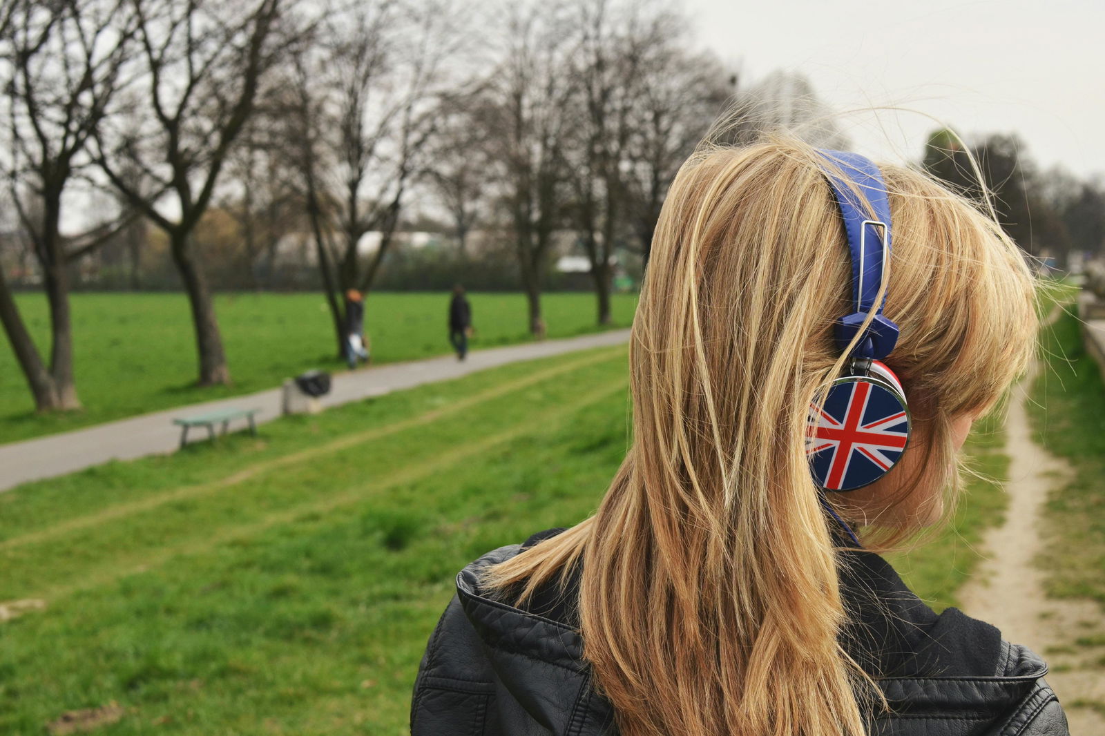 Eine junge Blondine hört etwas auf ihren Kopfhörern mit Union-Jack-Motiv an, während sie in der Natur durch den Park läuft