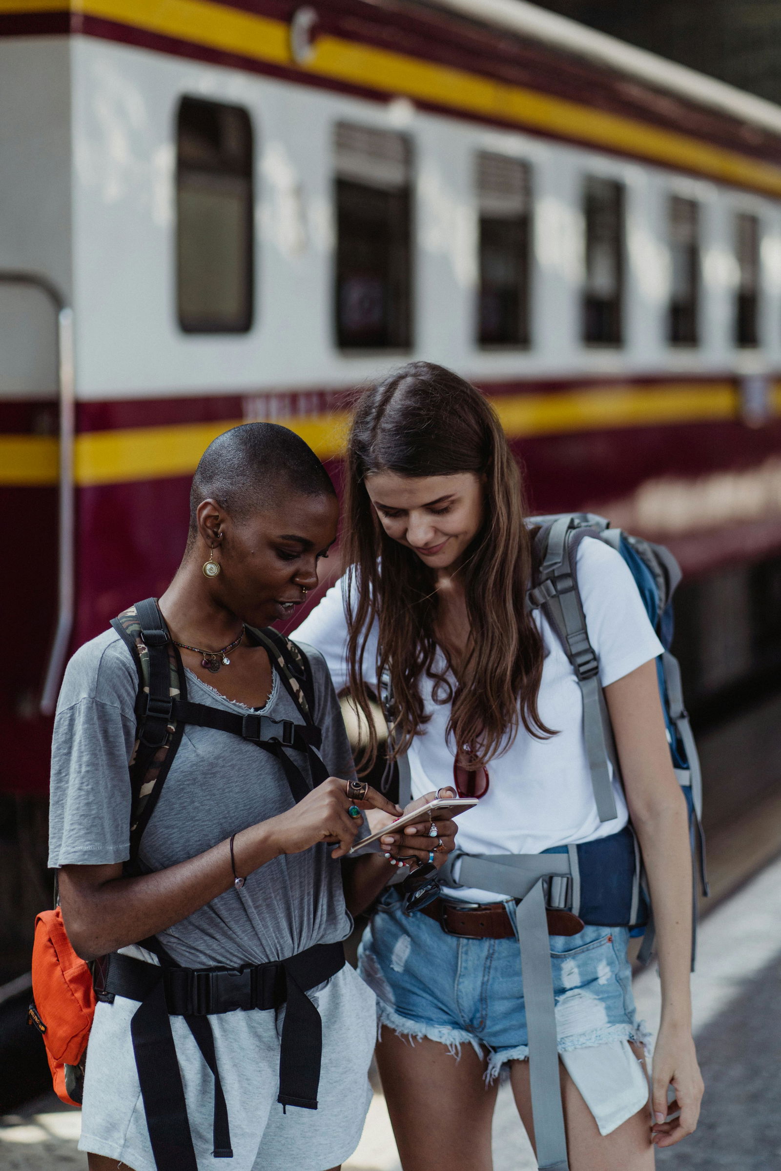 Zwei junge Reisende machen ein Gap Year und bereisen gemeinsam die Welt. Gerade stehen sie mit ihren Rucksäcken auf den Schultern vor einem Zug und suchen eine Zugverbindung raus. 