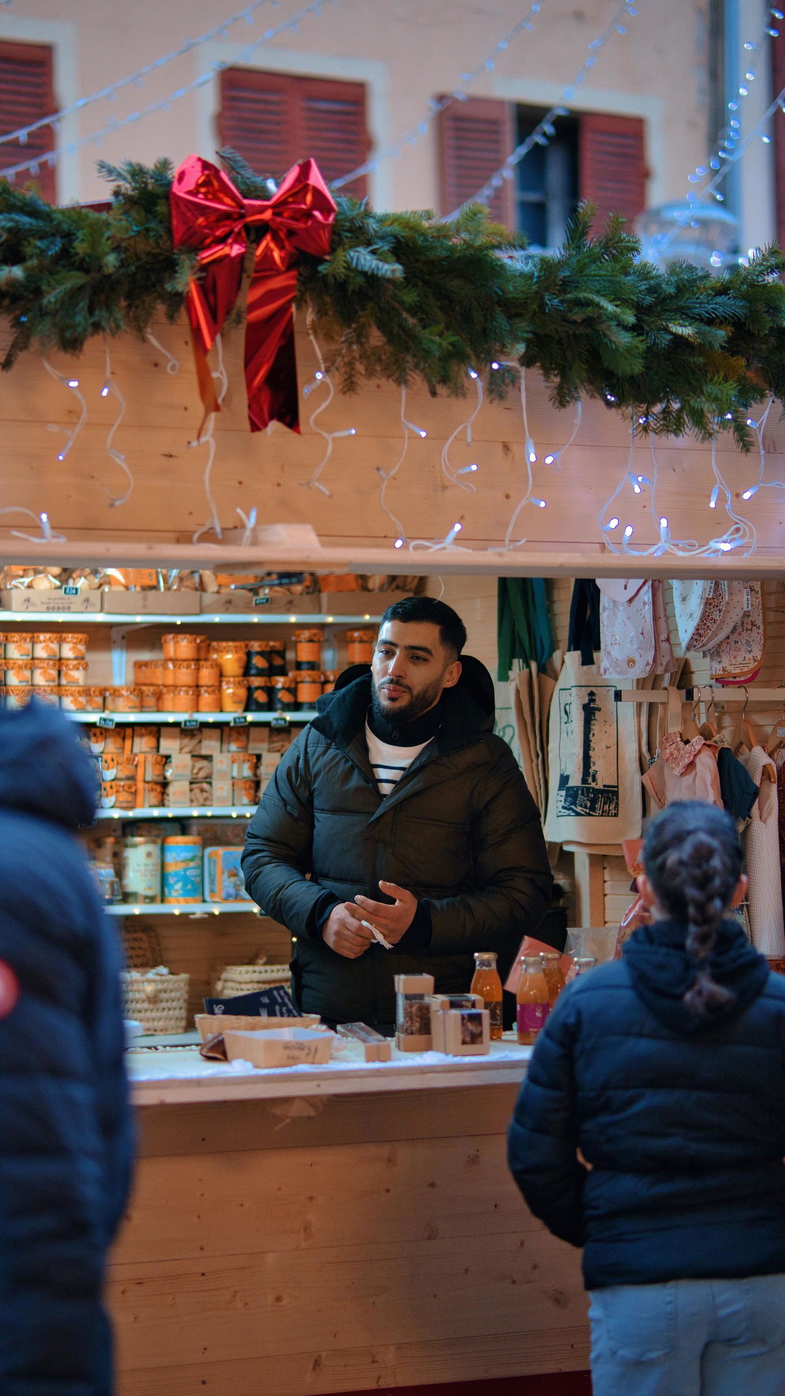Ein junger Mann steht in einer dicken Winterjacke in einem Weihnachtsmarkt-Verkaufshäuschen und verkauft Honig