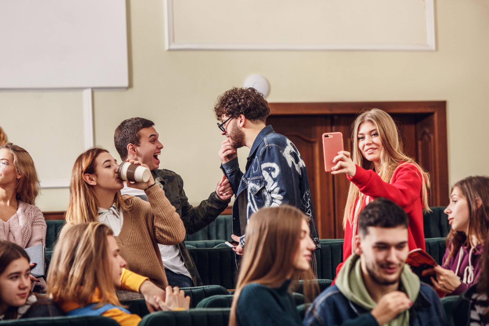 Studenten in den hinteren Hörsaal-Reihen unterhalten sich, lachen und trinken Kaffee. Eine Studentin schießt ein Selfie 