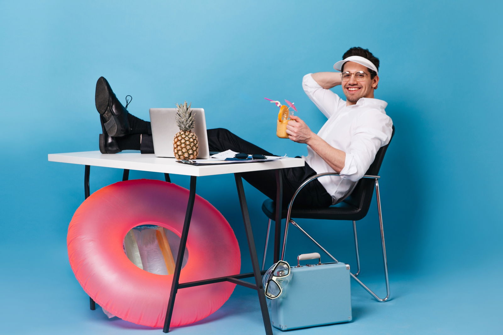 Ein Mann im Anzug sitzt an seinem Schreibtisch - die Beine auf den Tisch gelegt hält er einen Cocktail in der Hand und wähnt sich in Urlaubsstimmung 