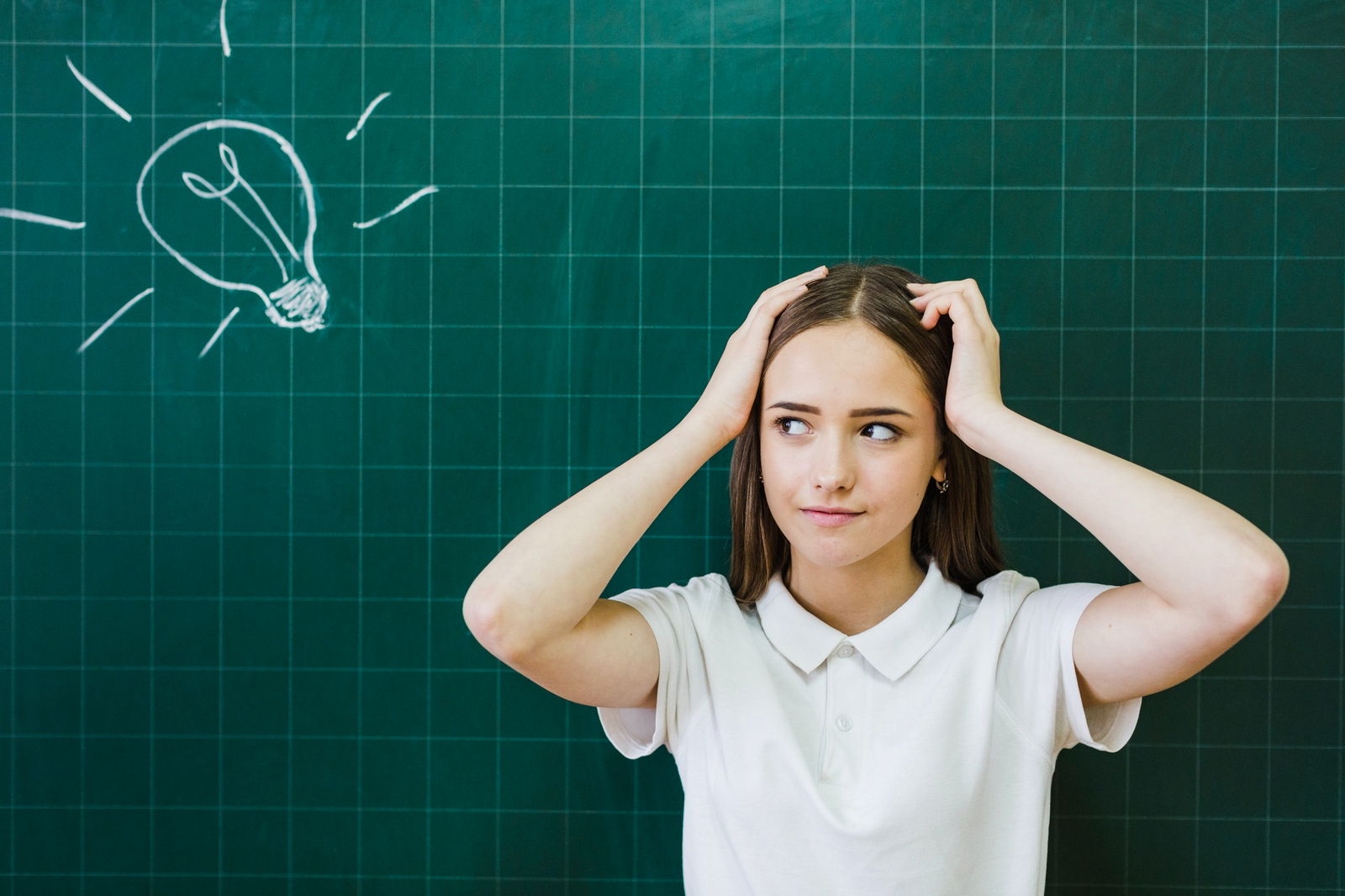 Eine junge Schülerin steht vor einer Tafel auf der mit Kreide eine Glühbirne aufgezeichnet ist - sie überlegt und hält die Hände über dem Kopf zusammen