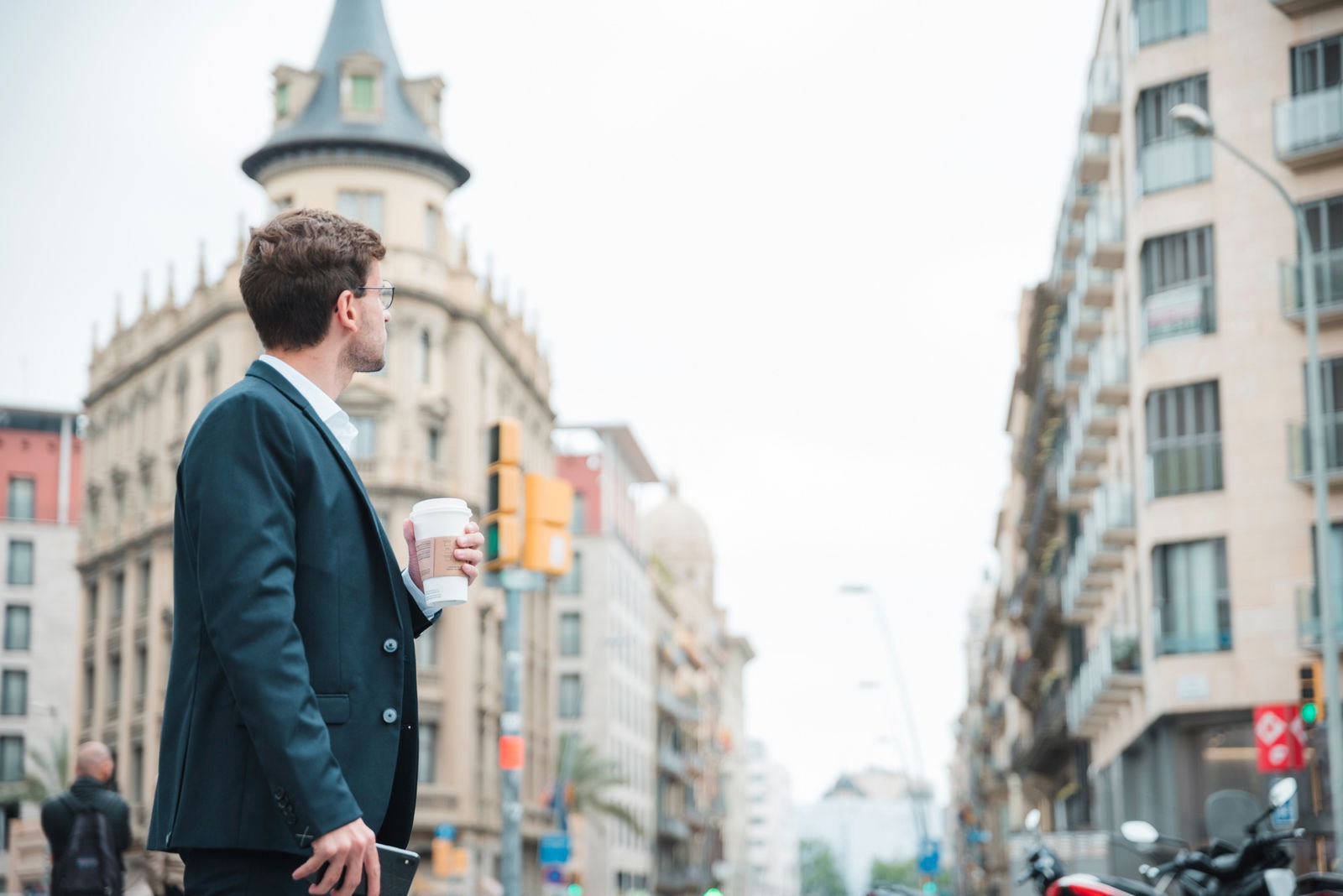 Ein Mann im Anzug schaut auf eine Häuserschlucht in einer Großstadt. In seiner Hand hält er einen Coffee-To-Go-Becher 