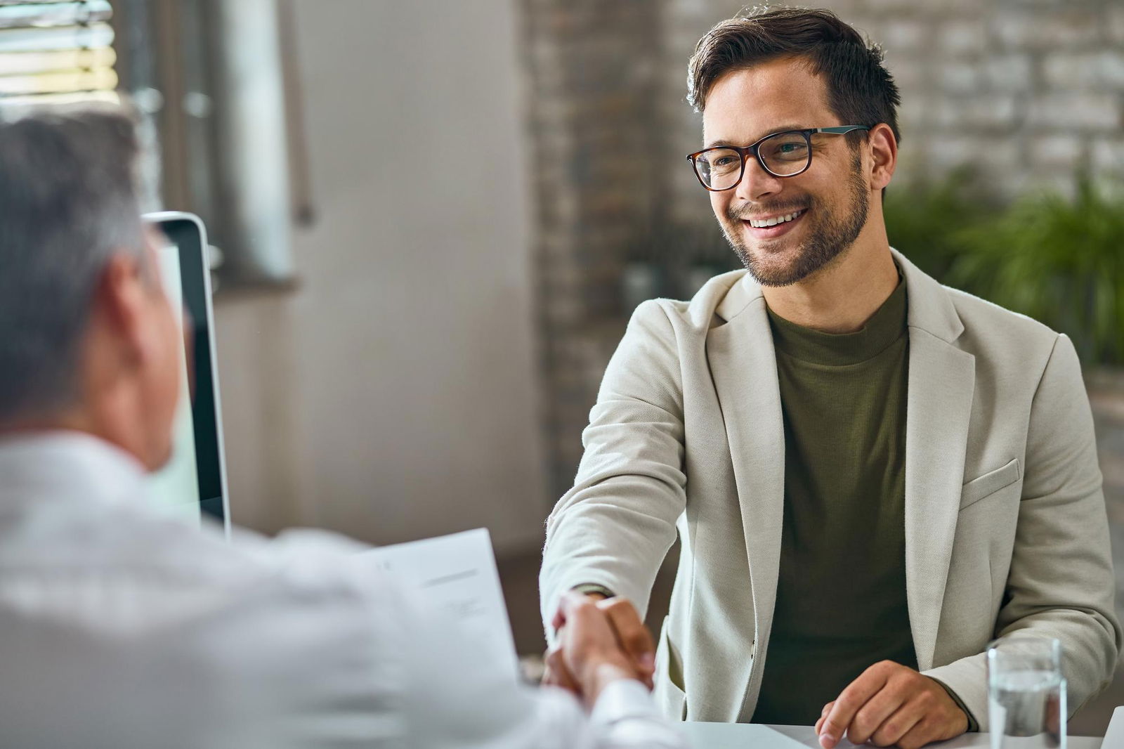 Ein junger Mann mit Brille sitzt einem Personaler gegenüber, der dessen Bewerbung in der Hand hält. Beide Männer schütteln sich die Hände und der Bewerber lacht. 