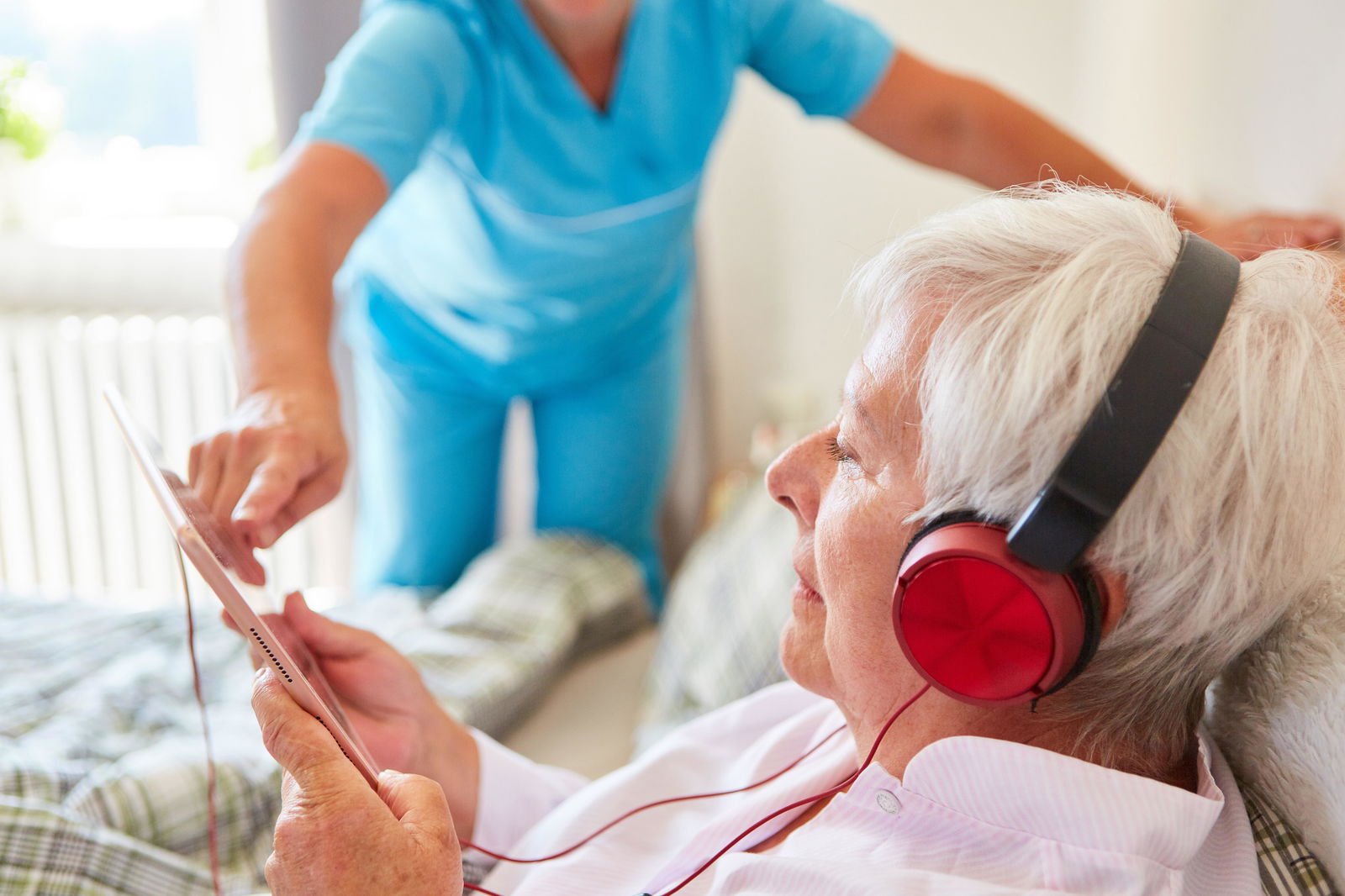 Eine alte Dame im Krankenhaus liegt mit Kopfhörern auf den Ohren im Bett und eine Pflegerin erklärt ihr etwas auf dem Tablet