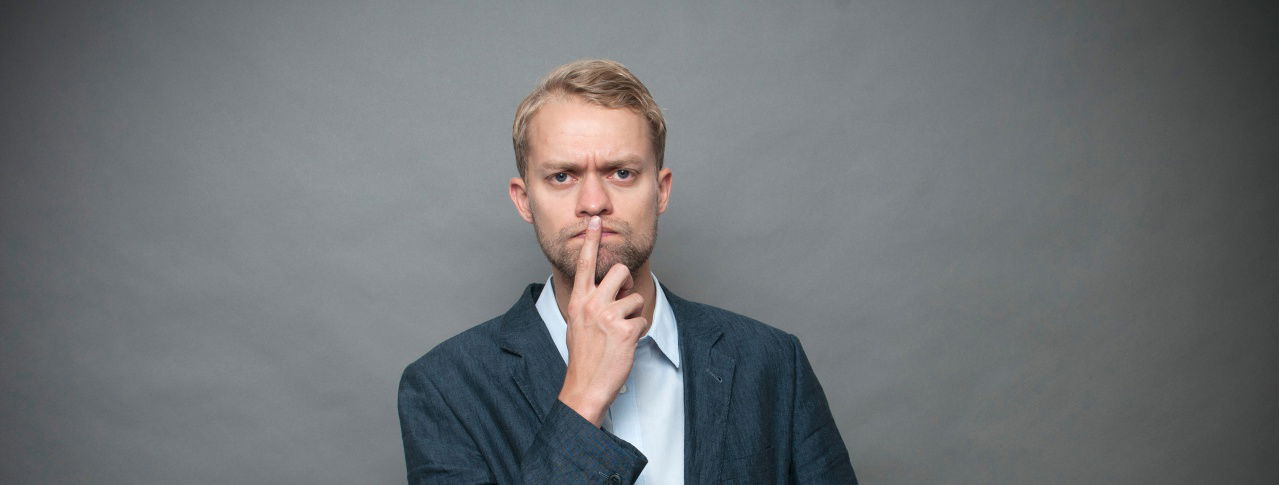 Nachdenklicher Mann vor grauen Wand, Zeigefinger vorm Mund und schaut in Kamera