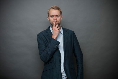 Nachdenklicher Mann vor grauen Wand, Zeigefinger vorm Mund und schaut in Kamera