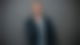 Nachdenklicher Mann vor grauen Wand, Zeigefinger vorm Mund und schaut in Kamera