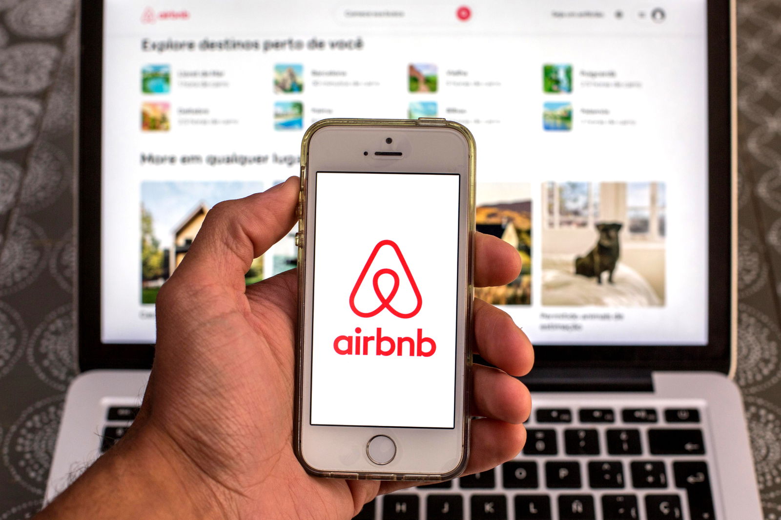 Ein Smartphone zeigt das Airbnb Logo, im Hintergrund ist ein Notebook mit Urlaubszielen zu sehen