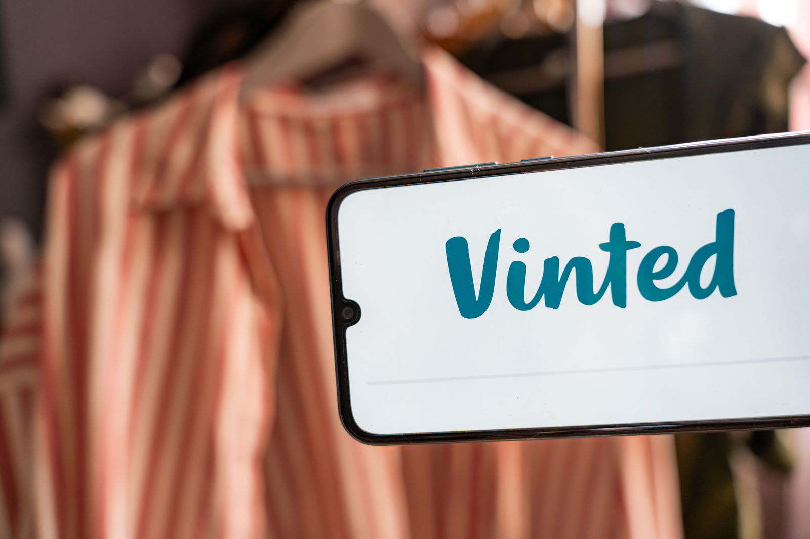 Auf einem Handy-Display ist das Wort Vinted zu lesen - der Name einer Verkaufsplattform für Second Hand Mode. Im Hintergrund ist ein Kleidungsstück an einem Kleiderbügel zu sehen.