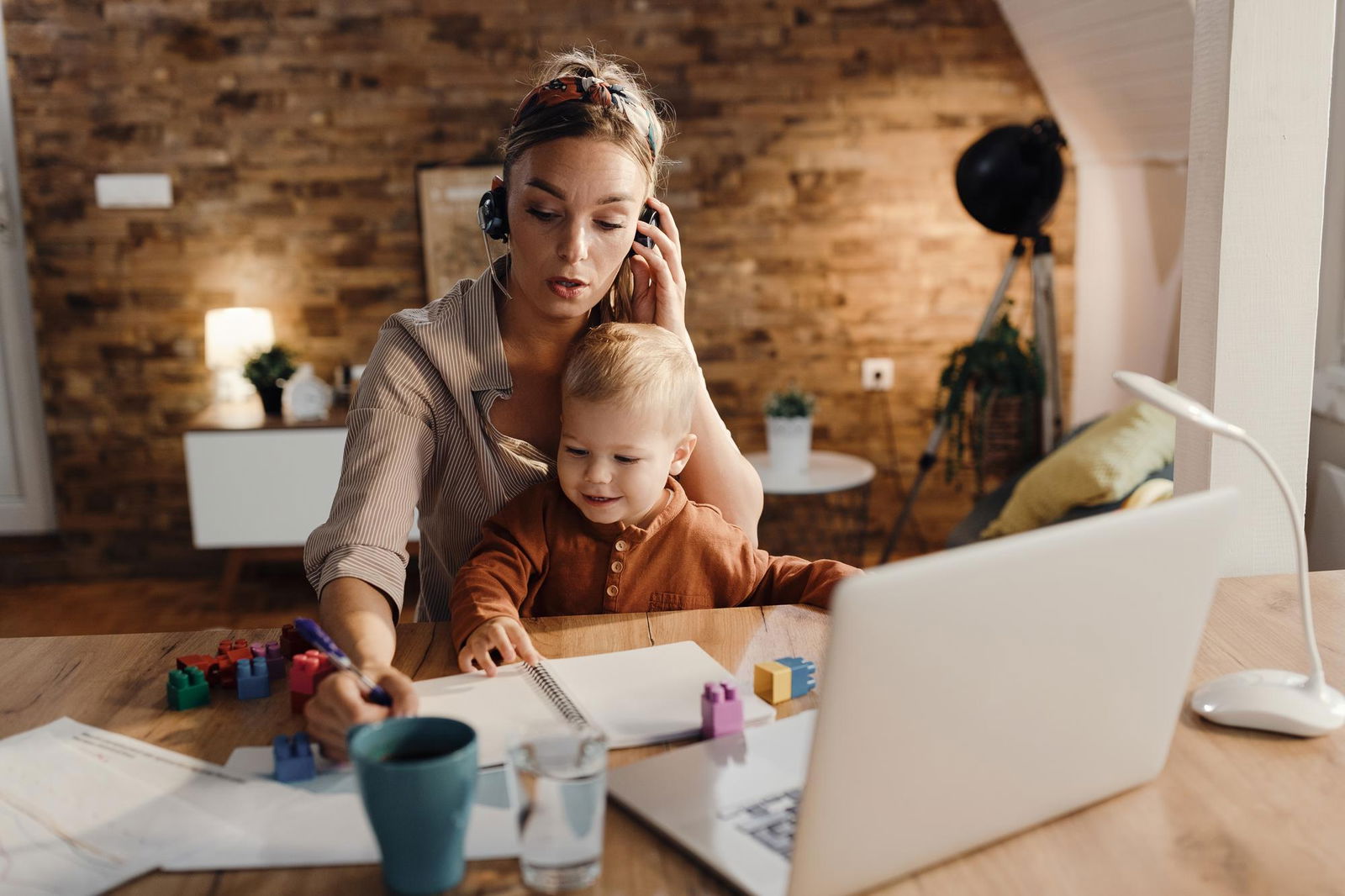 Berufstätige Mutter mittleren Alters mit Kleinkind auf dem Schoß macht sich während eines Videoanrufs im Homeoffice Notizen. Um sie herum liegen Duplo-Steine mit dem der kleine Junge spielt