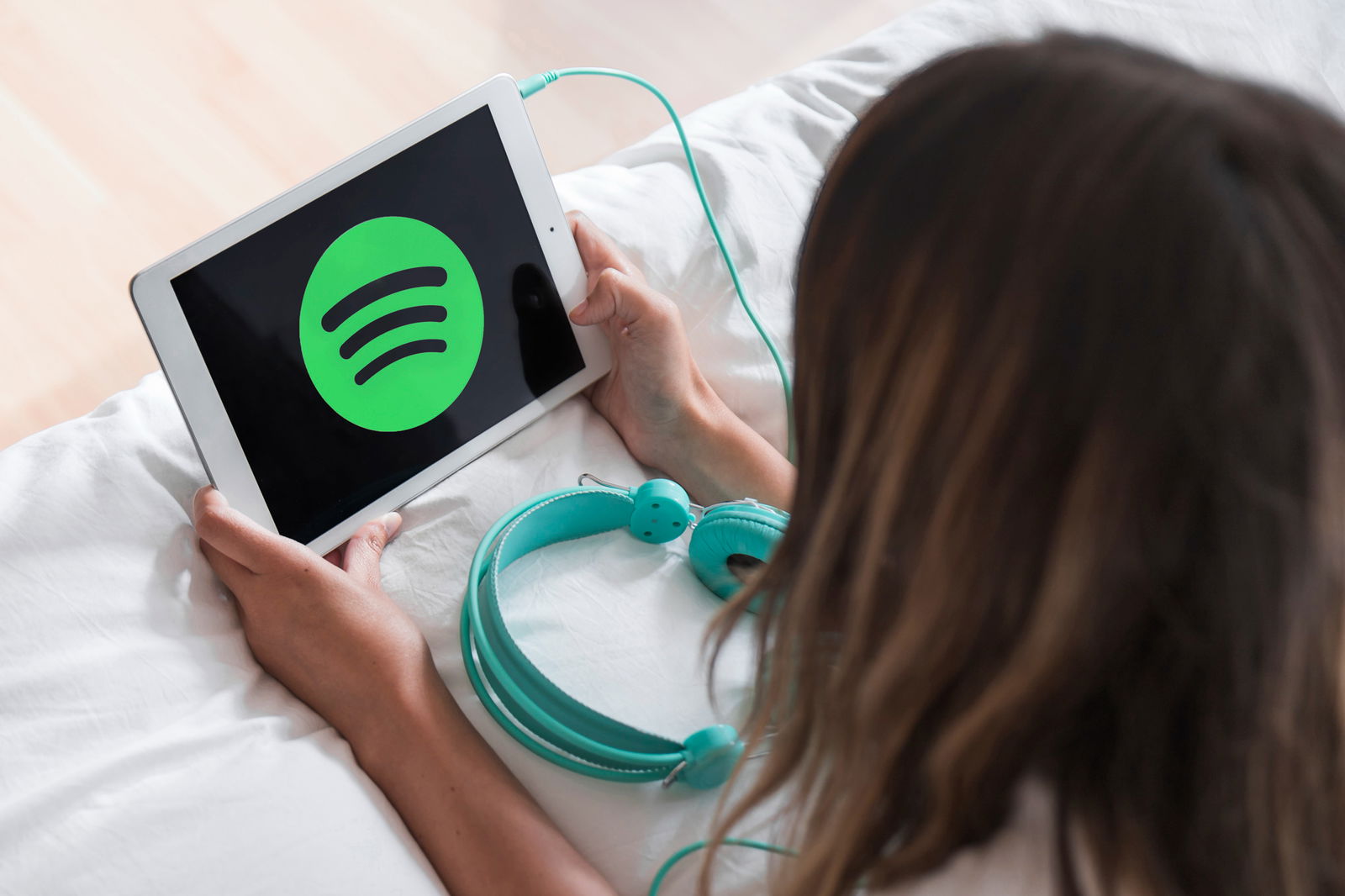 Eine junge Frau liegt bäuchlings auf dem Bett. Vor sich hält sie ein Tablet auf dem das Logo des Audio-Streaming-Diensts Spotify abgebildet ist