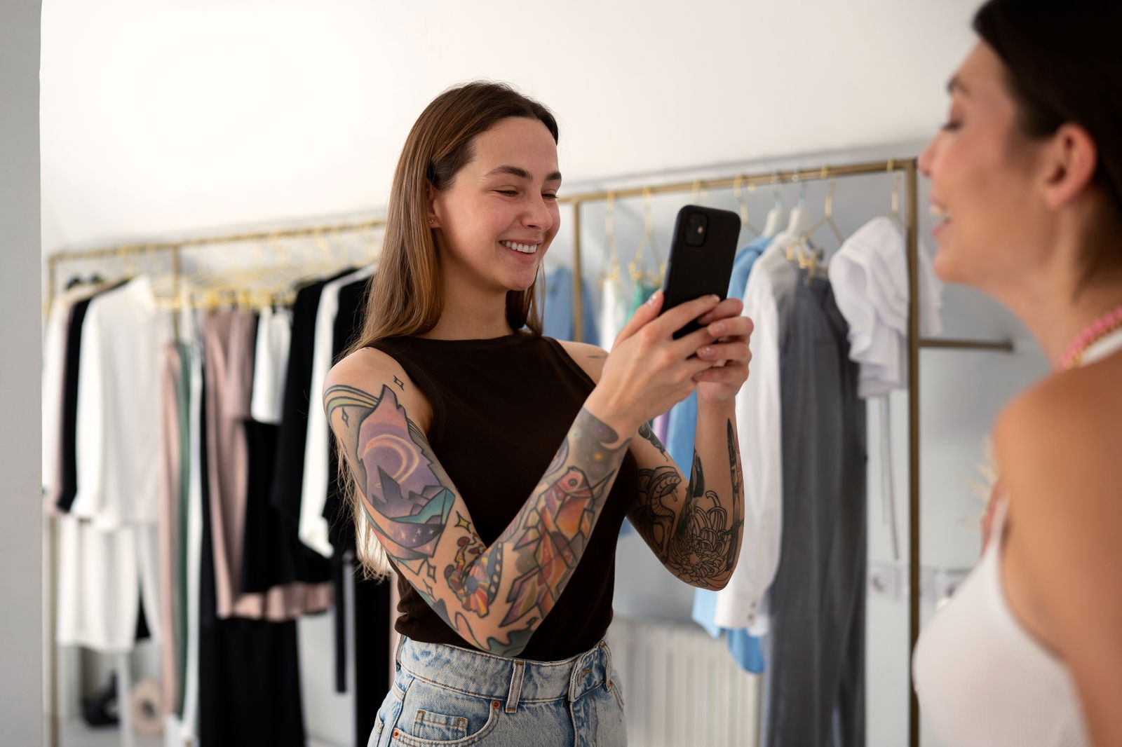 Eine junge, tattowierte Frau macht in einem Bekleidungsgeschäft ein Foto ihrer Freundin, die gerade ein Oberteil anprobiert. Beide lachen
