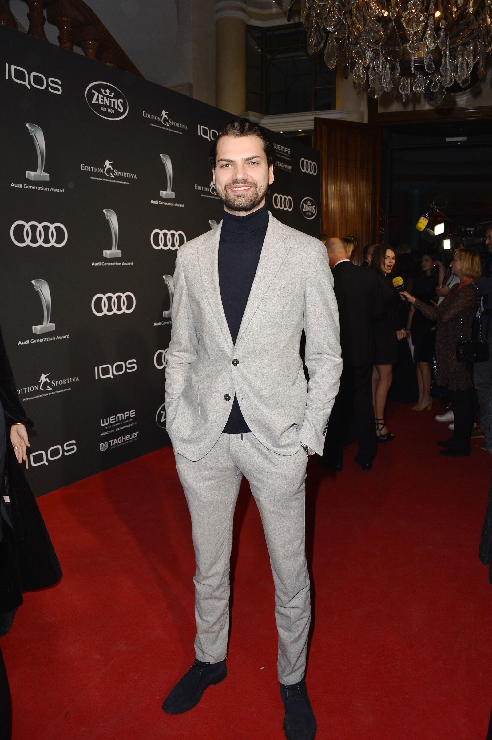 der_ehemalige_schauspieler_jimmi_blue_ochsenknecht_steht_in_einem_grauen_anzug_und_schwarzen_rollkragen-pullover_auf_einem_roten_teppich. Beide Hände in den Hosentaschen. Er lacht.jpg