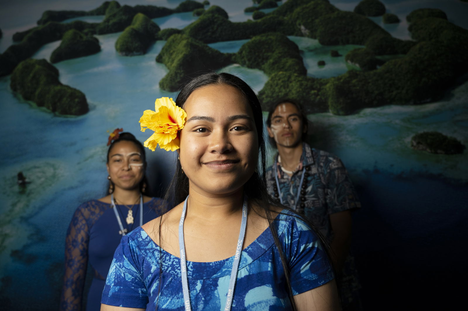 Drei junge Bewohnerinnen des Inselstaates Tuvalu stehen vor einem Hintergrund mit türkisblauem Wasser und grünen Inseln. Die Frau im Vordergrund trägt ein blaues Kleid und eine gelbe Blume im Haar. Alle tragen Namensbänder mit der Aufschrift „UNFCCC“