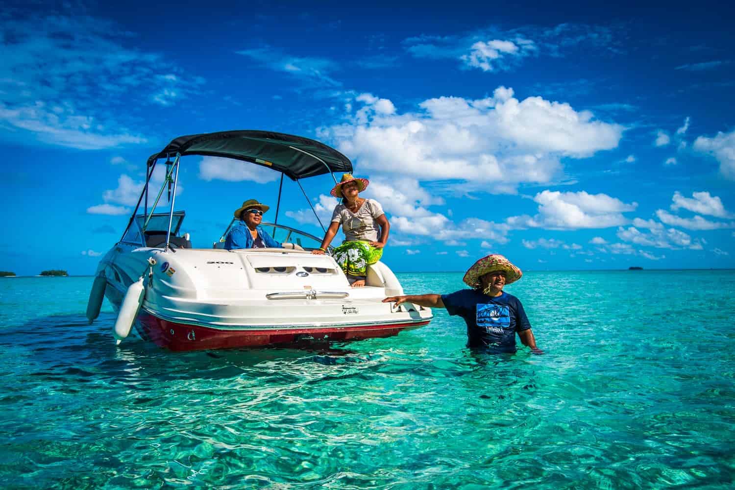 Drei Personen aus Tuvalu tragen Strohhüte und bunte Kleidung. Zwei von ihnen sitzen auf einem weißen Motorboot, eine Person steht lachend im türkisblauen, flachen Wasser neben dem Boot. 