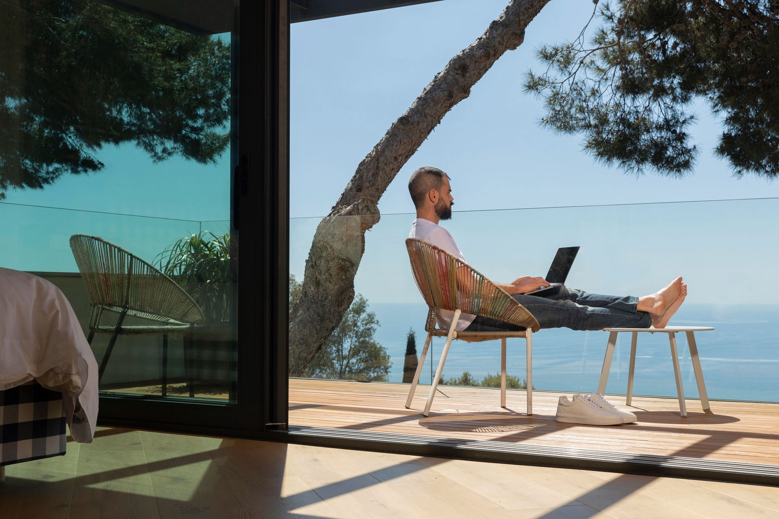 Ein Mann sitzt barfuß mit Laptop auf einer Terrasse am Meer – statt zu entspannen, arbeitet er im Urlaub.