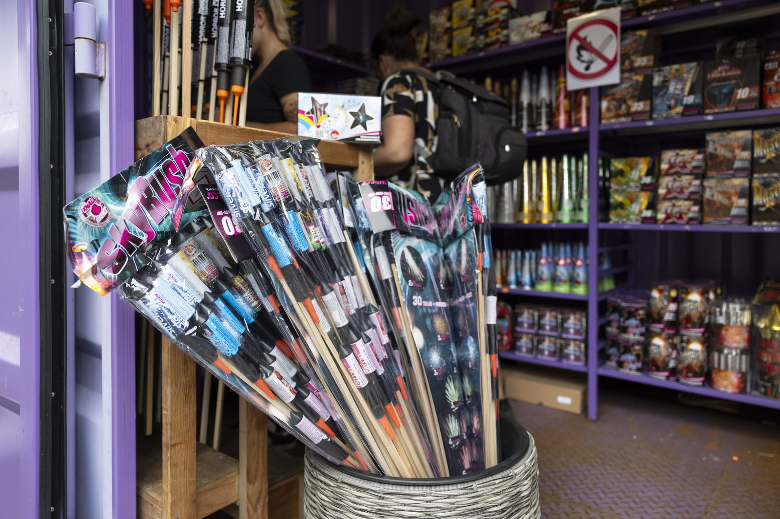 Ein Verkaufsstand für Silvesterfeuerwerk mit bunten Raketenbündeln und Regalen voller Pyrotechnik. Zwei Personen stehen an der Theke