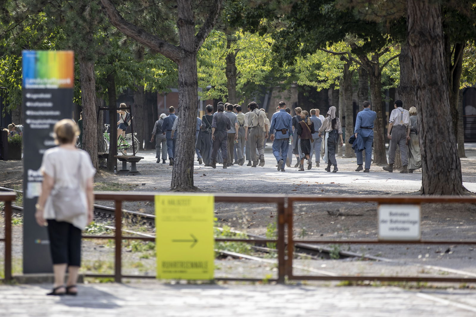 Eine Gruppe von Kompars:innen in historisch anmutender Kleidung geht auf einem von Bäumen gesäumten Weg im Landschaftspark Duisburg. Sie nehmen an Dreharbeiten zum neuen Film „Die Tribute von Panem: Das Lied von Vogel und Schlange“ teil. 