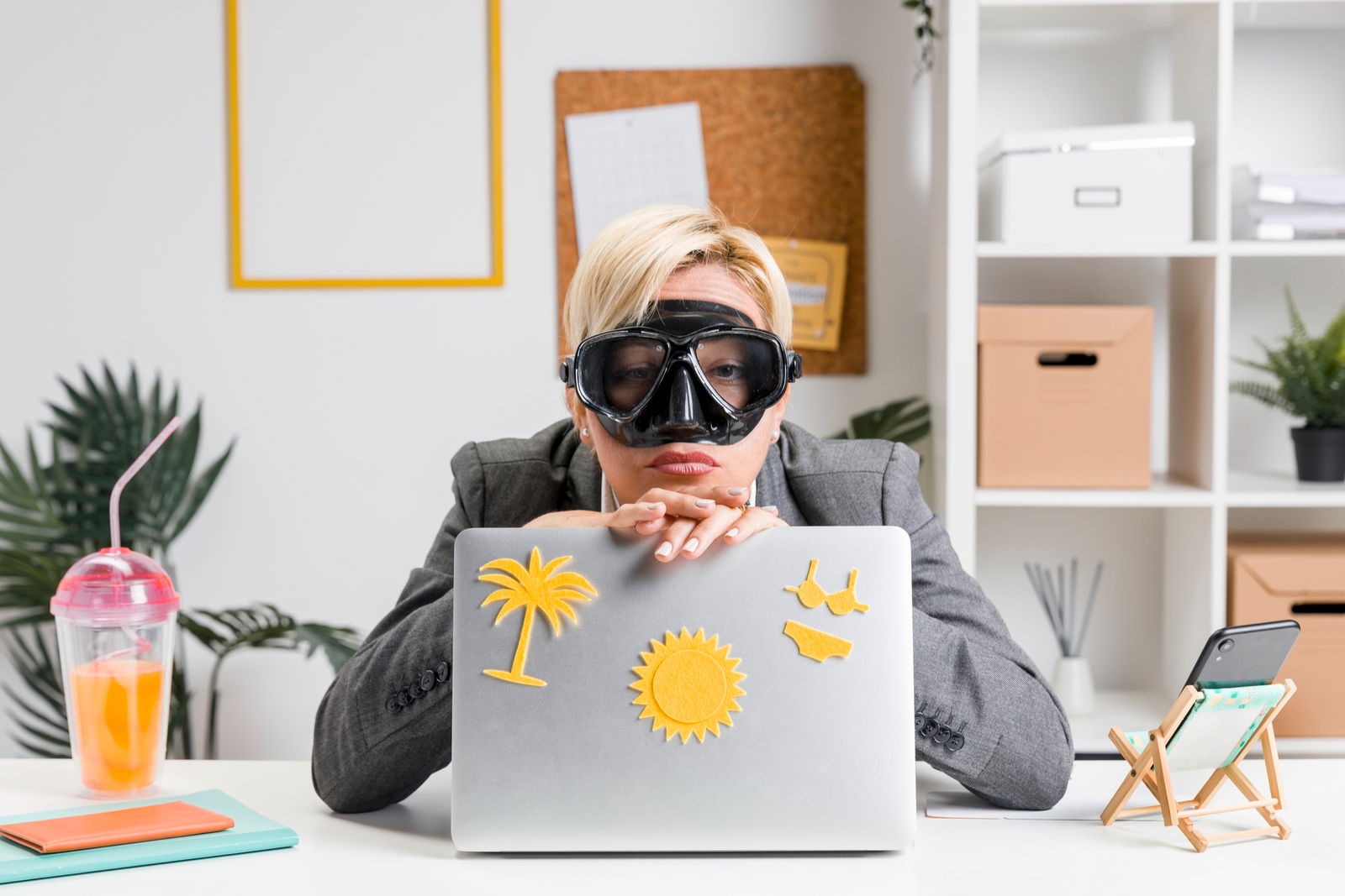 Eine Frau sitzt im Büro, trägt eine Tauchermaske und stützt sich müde auf ihren Laptop – dekoriert mit Sommermotiven. Der Urlaub ist sichtbar fern, die Erschöpfung nah.
