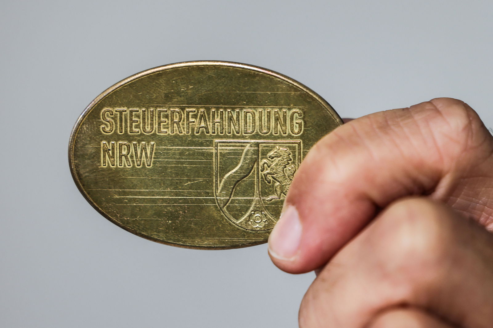 Eine Hand hält eine goldfarbene Dienstmarke mit der Aufschrift „Steuerfahndung NRW“ und dem Wappen von Nordrhein-Westfalen.