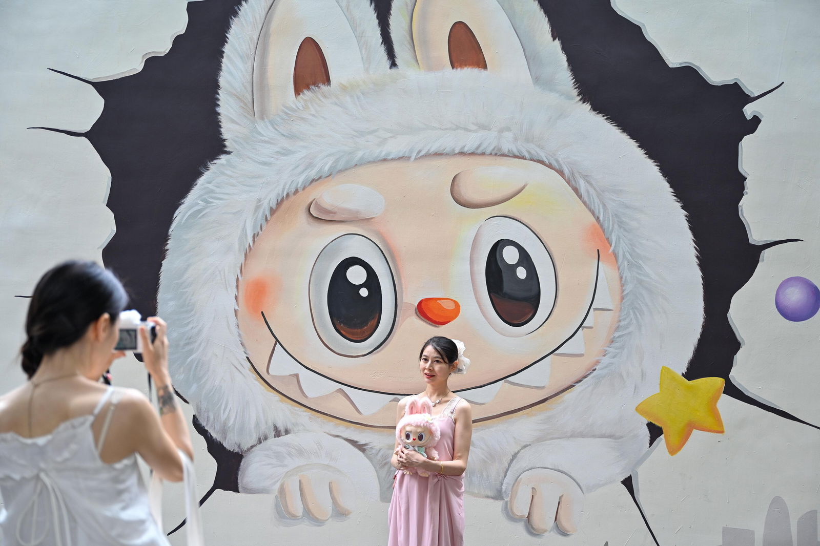 Eine junge Frau in rosa Kleid und weißem Haarschmuck steht vor einem großen Wandbild der Comicfigur „Labubu“. Die Figur hat große, runde Augen, ein breites Grinsen mit spitzen Zähnen und trägt ein weißes Fell