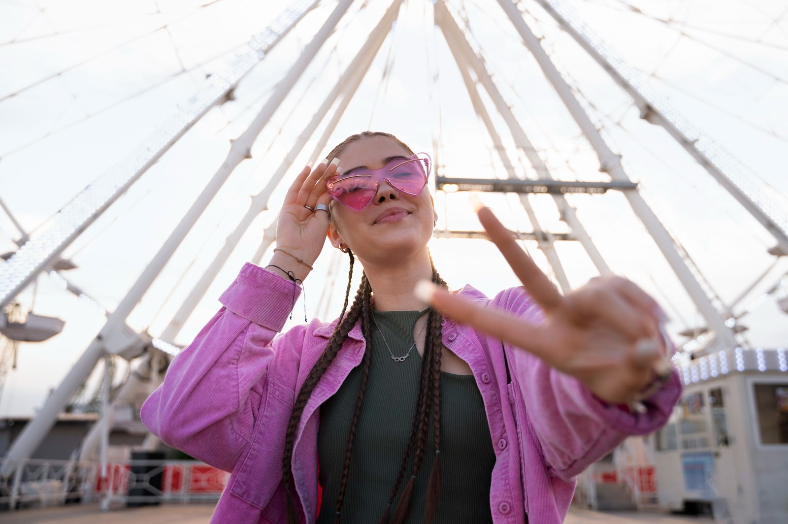 Eine junge Frau mit Zöpfen steht vor einem Riesenrad, trägt eine pinke Herz-Sonnenbrille und zeigt ein Peace-Zeichen zur Kamera.