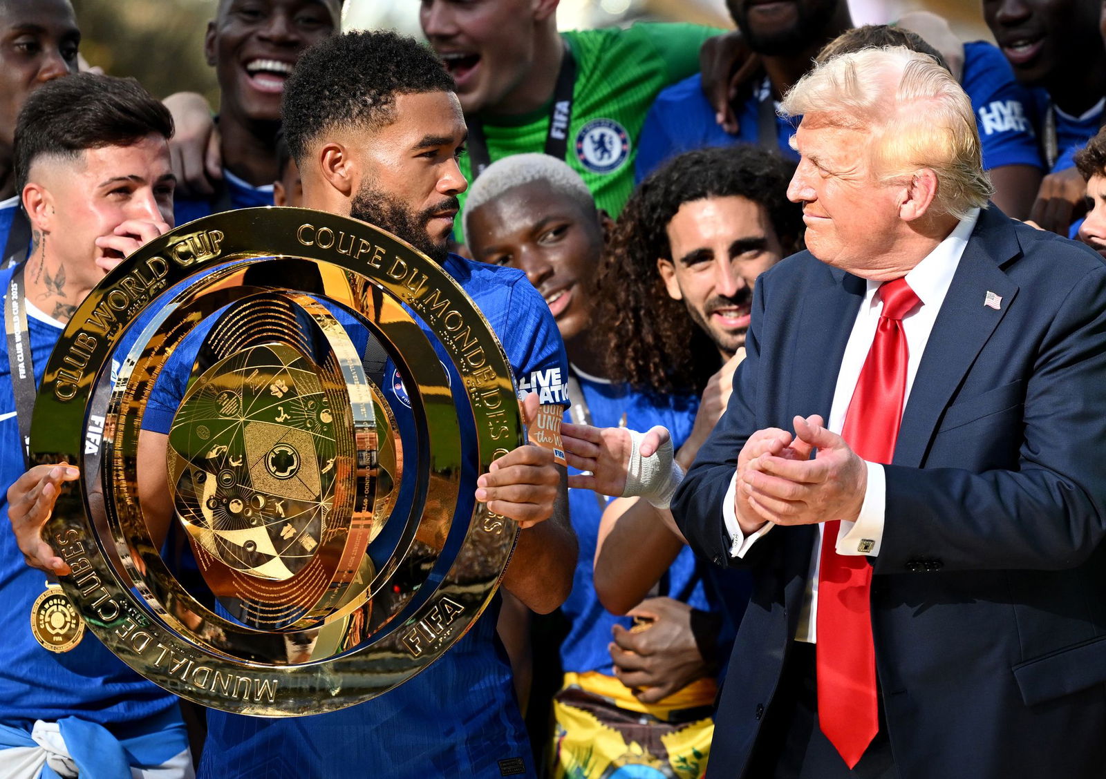 Chelseas Kapitän Reece James (l/vorn) hält auf dem Siegerpodest die Trophäe in den Händen und schaut irritiert zu US-Präsident Donald Trump.