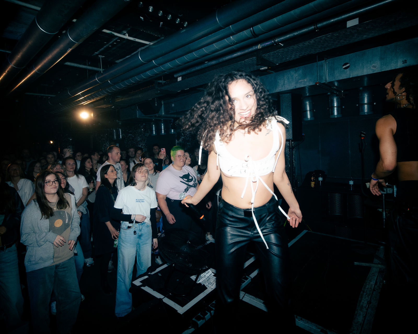 Eine junge Frau mit braunen lockigen Haaren hüpft glücklich strahlend über eine Bühne. Hinter ihr stehen Fans und schauen sie an. Die Frau trägt eine Lederhose und ein weißes Oberteil.