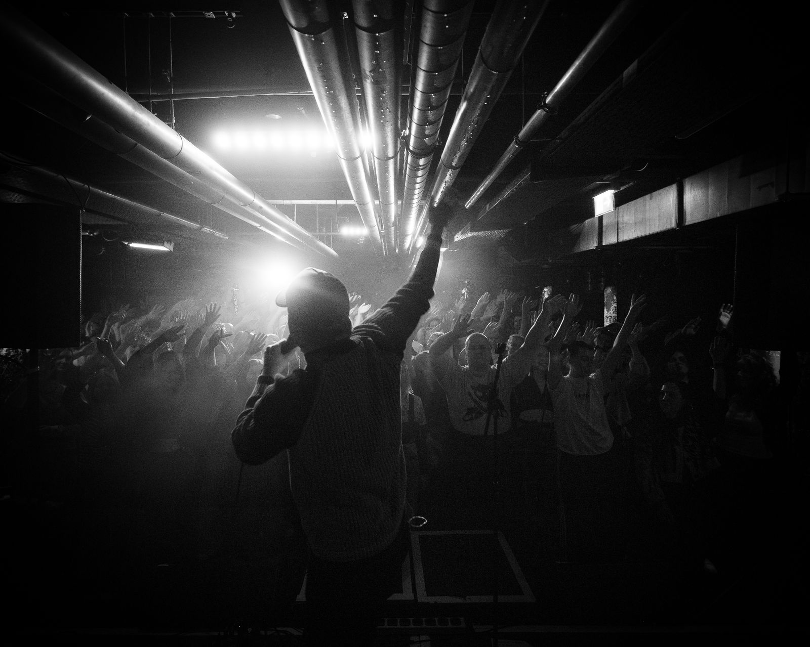 Ein Mann steht mit dem Rücken zur Kamera auf der Bühne und singt für ein Publikum in einem Club. Er streck den einen Arm und hält in der anderen Hand ein Mikrofon. Das Bild ist in schwarz-weiß aufgenommen.