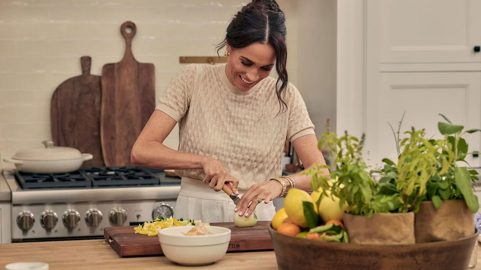 Meghan Markle in Loro Piana steht in ihrer Küche vor ihrem Herd und schneidet im Rahmen ihrer Netlflix-Serie eine Zwiebel. Sie lacht dabei