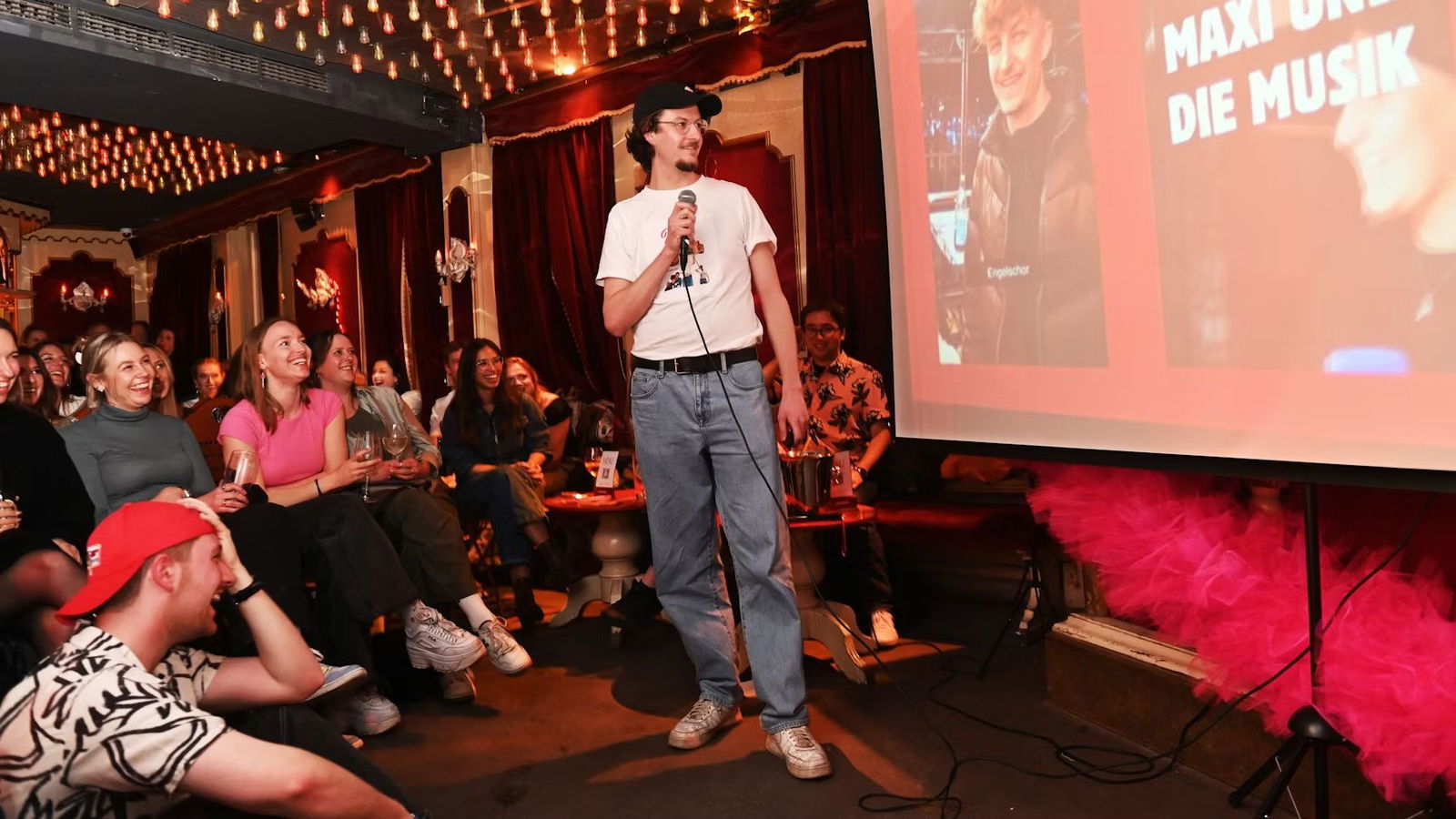 Ein junger Mann steht in einer Bar vor einer Leinwand auf der er eine Power-Point-Präsentation abhält. Auf der Präsentation ist ein Freund von ihm zu sehen, der Single ist. Der Mann mit dem Mikrofon in der Hand möchte ihn für Dating vorstellen.