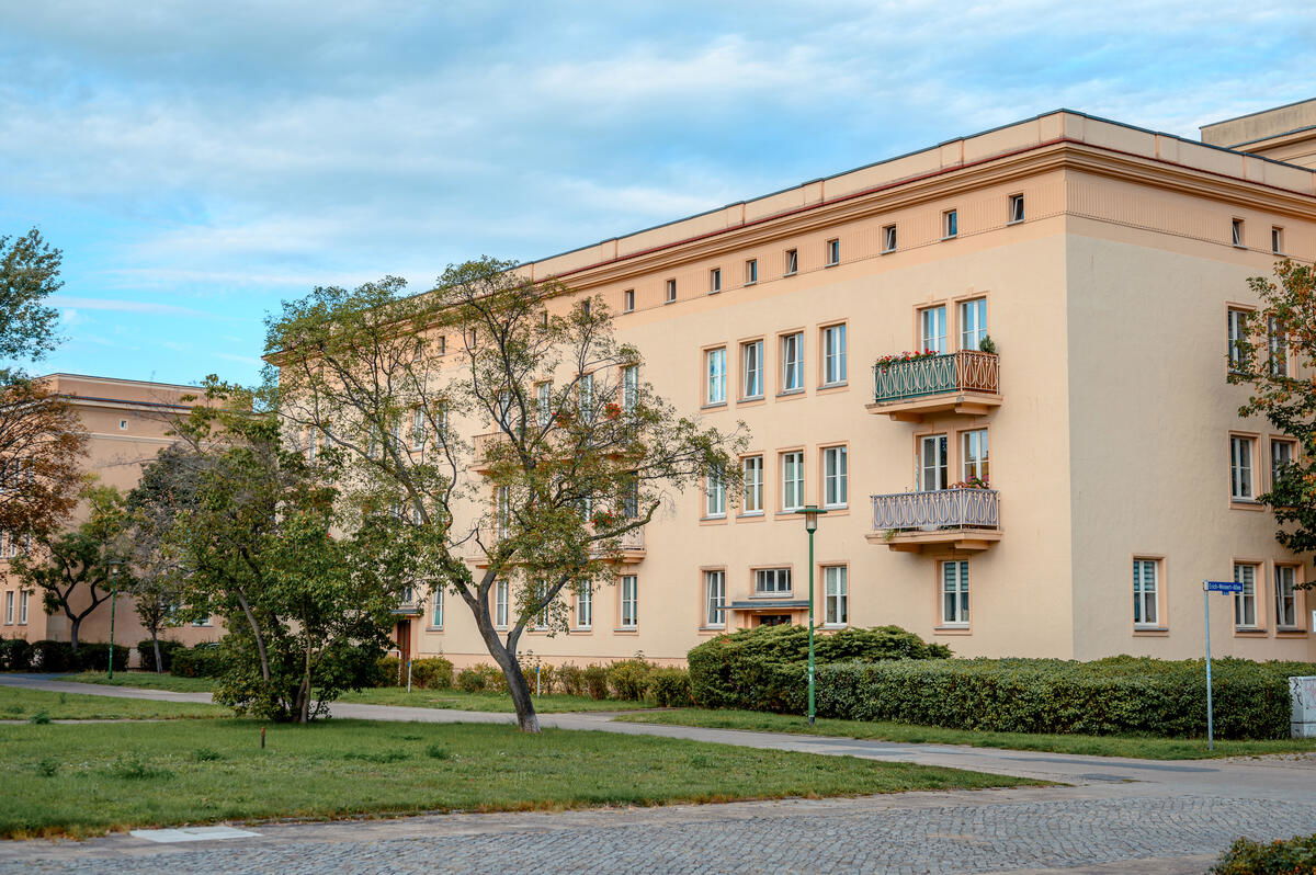 Ein helles Wohngebäude im Stil der Planstadt Eisenhüttenstadt mit symmetrischer Fassade, Balkonen und gepflegtem Grünstreifen im Vordergrund. Der Himmel ist leicht bewölkt, die Umgebung wirkt ruhig und geordnet.
