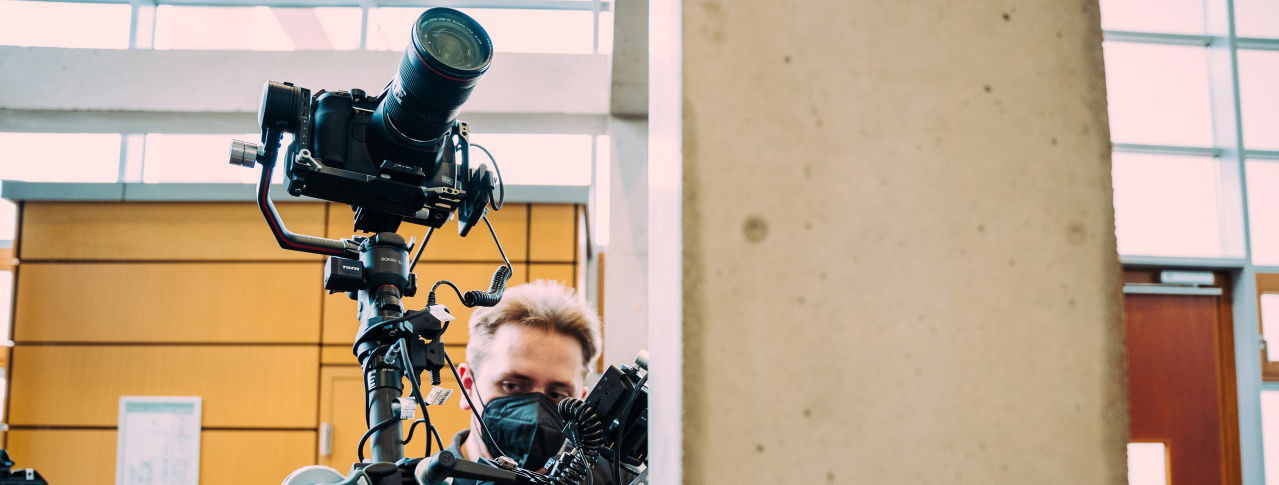 Kamera mit Stativ und ein Mann mit Maske steht dahinter