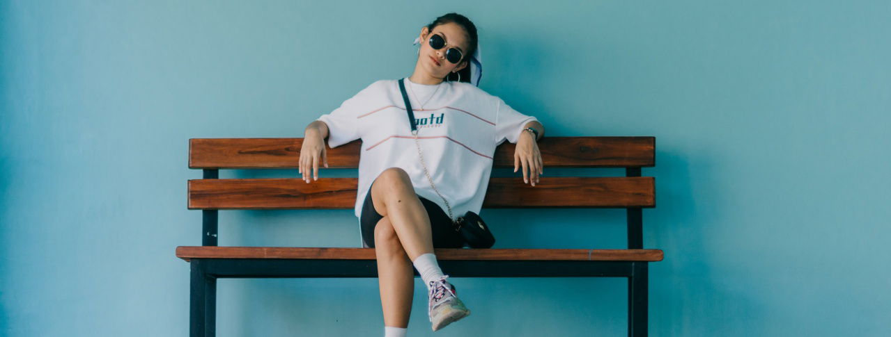 Mädchen sitzt cool und locker mit einer Sonnenbrille und überkreuzten Füßen auf einer Bank