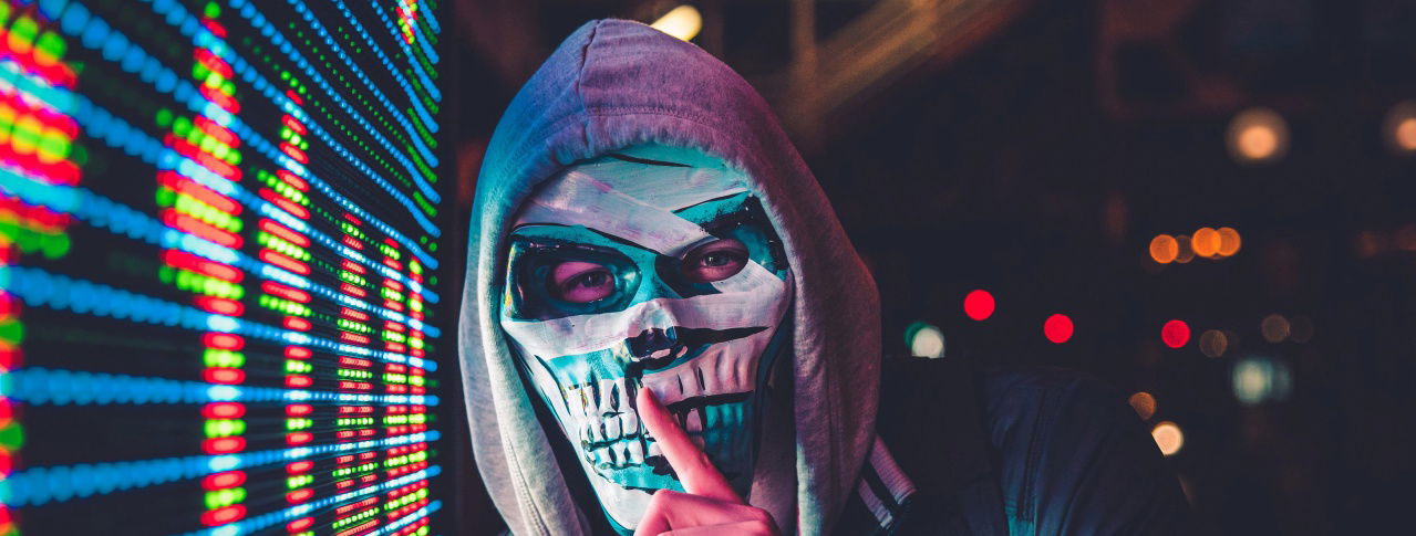 Mann mit Totenkopfmaske hält Finger vor Mund und hat Kapuze auf