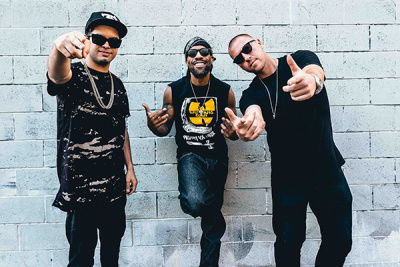 Drei Männer sind relativ schwarz gekleidet und zeigen mit ihren Händen in die Kamera, zudem tragen sie alle eine Sonnenbrille