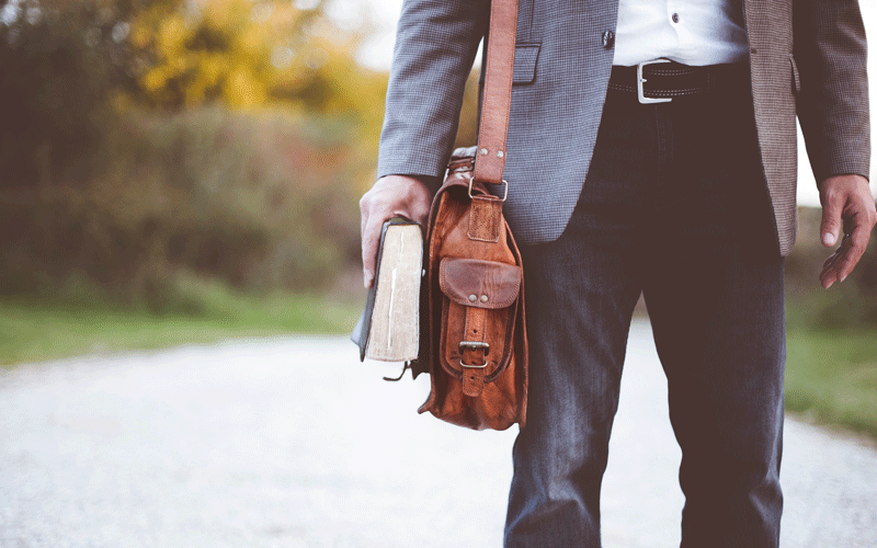 Mann ist schick angezogen, um seine Schulter hat er seine Tasche hängen und hält ein Buch in der Hand 