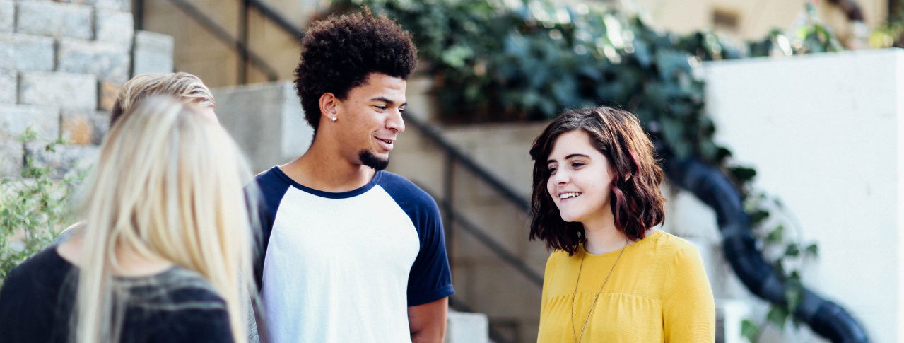 Ein Mann unterhält sich mit zwei Frauen, die eine steht mit dem Rücken zur Kamera und die andere Frau hat ein gelbes Oberteil an und braune kurze Harre und lächelt