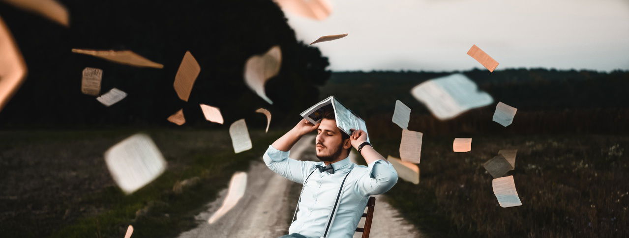 Mann sitzt auf einem Stuhl auf dem Feld und hebt ein Buch über sich und um ihn herum fliegen viele Blätter 